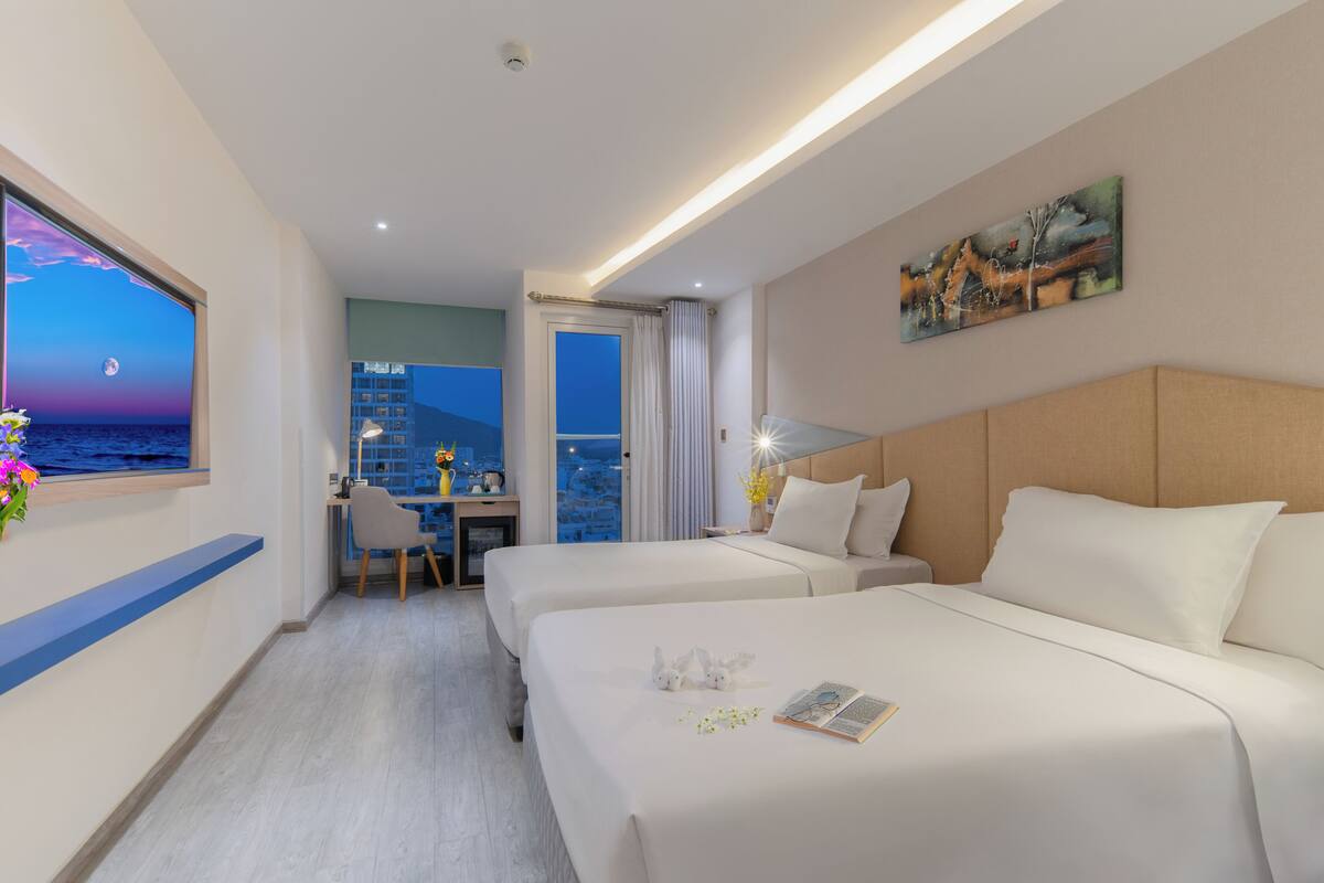 A hotel room with two beds, a desk with a chair, a window with a view of the city at night, and a painting on the wall.