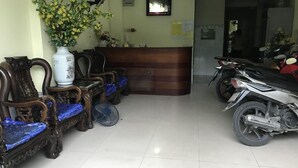 A reception area with a reception desk, several wooden chairs with blue cushions, a white vase with flowers, and a motorcycle parked to the right.
