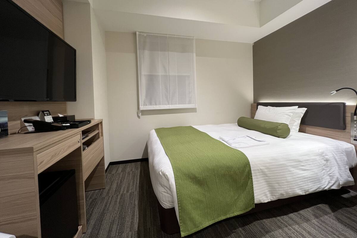 Una habitación de hotel con cama, escritorio, televisión y ventana con cortina translúcida.