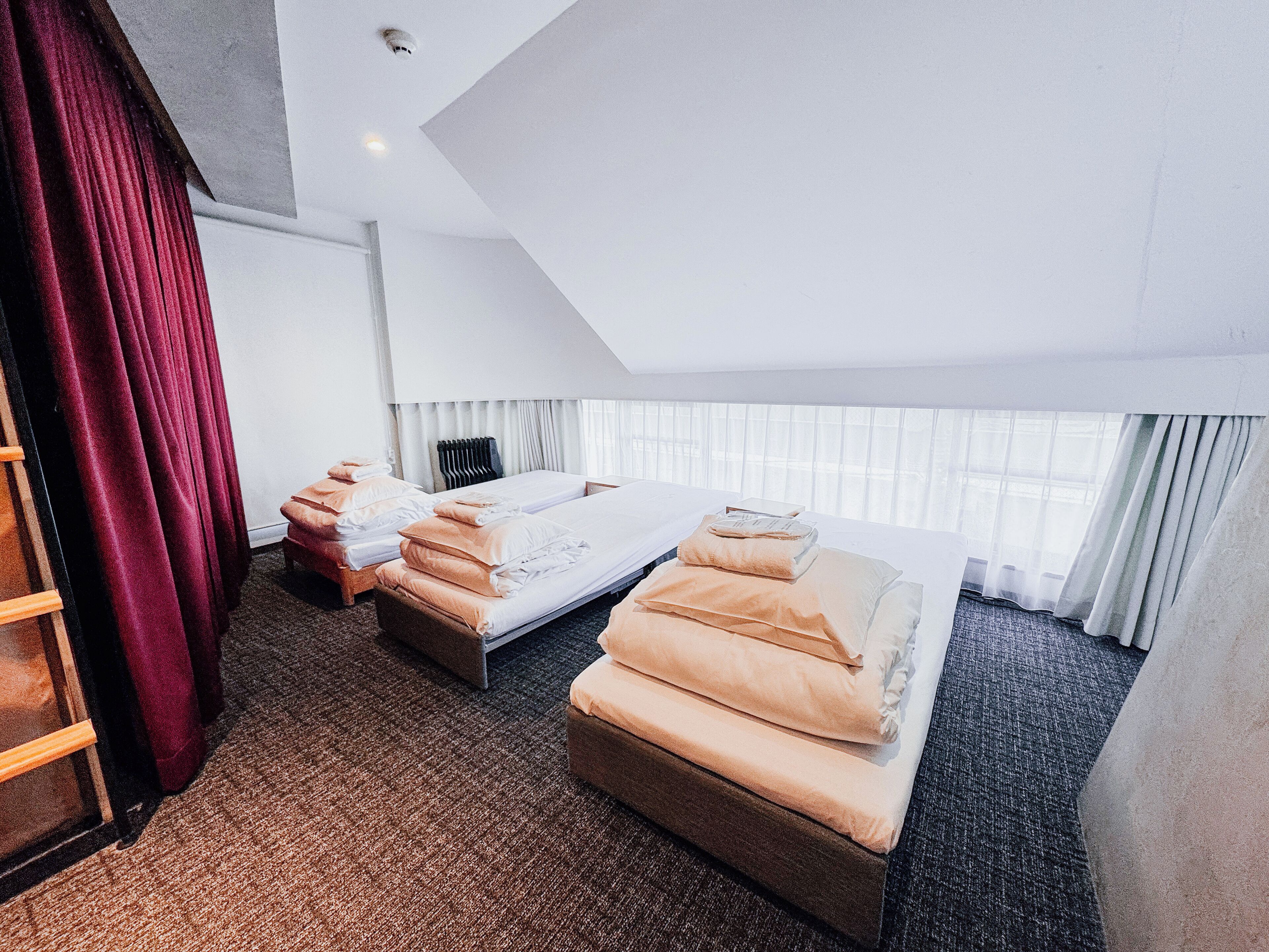 Une chambre d’hôtel avec deux lits, une grande fenêtre avec des rideaux légers, un rideau rouge sur le côté gauche et un sol recouvert de moquette.
