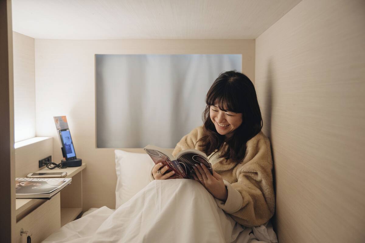 A person sitting on a bed in a room with a bedside table holding a book and a phone.