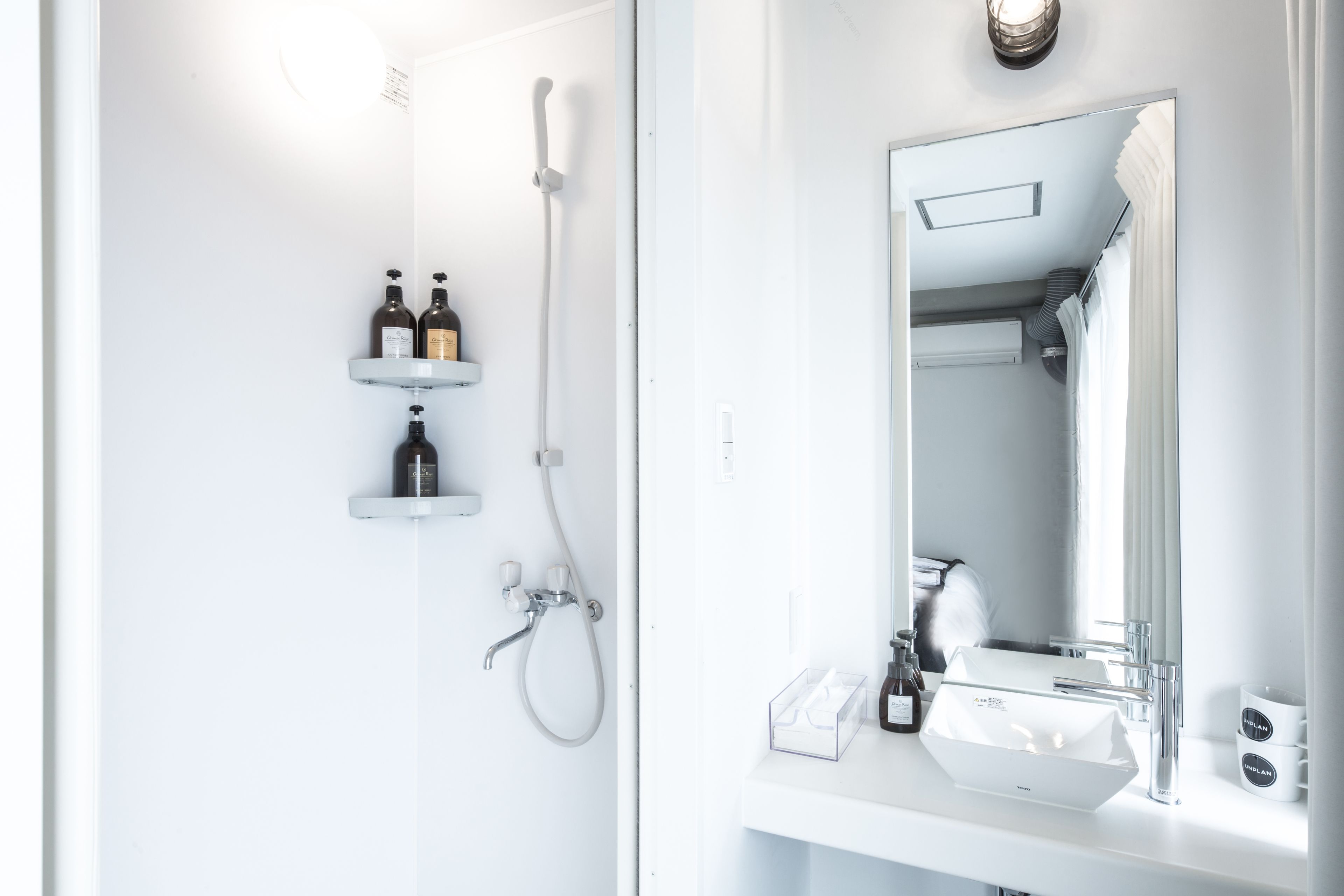 A modern bathroom with a white sink, a mirror, and a shower. There are two bottles on a shelf above the sink.