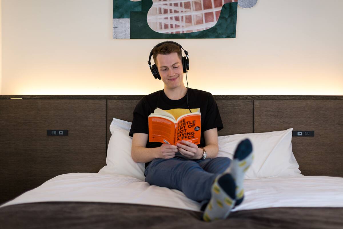 A person sitting on a bed with headphones, reading a book.