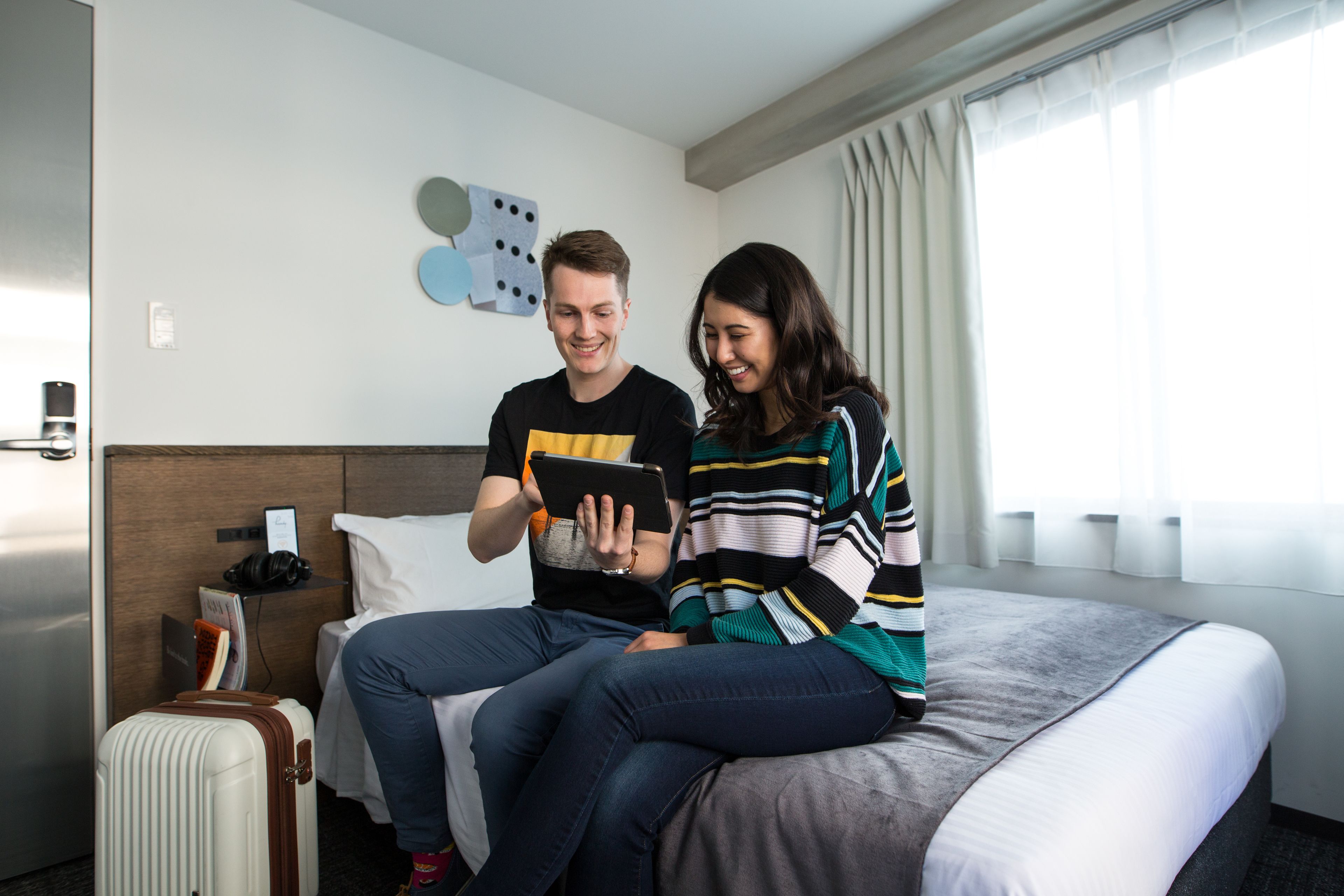 A hotel room with a bed, a suitcase, and a wall-mounted shelf with a book and headphones.