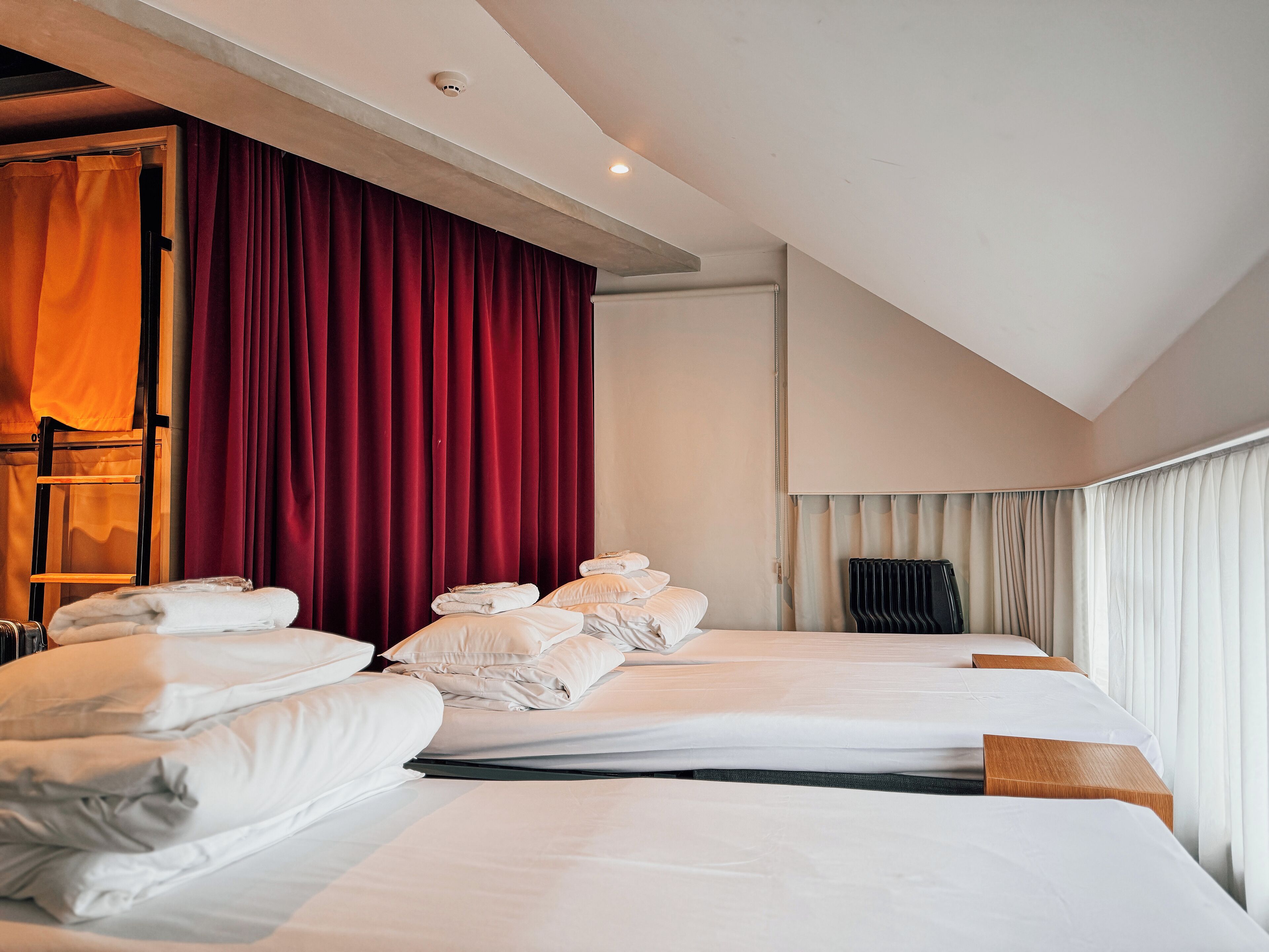 Une chambre d’hôtel avec deux lits, des rideaux rouges, du linge de lit blanc, un radiateur et un détecteur de fumée fixé au plafond.
