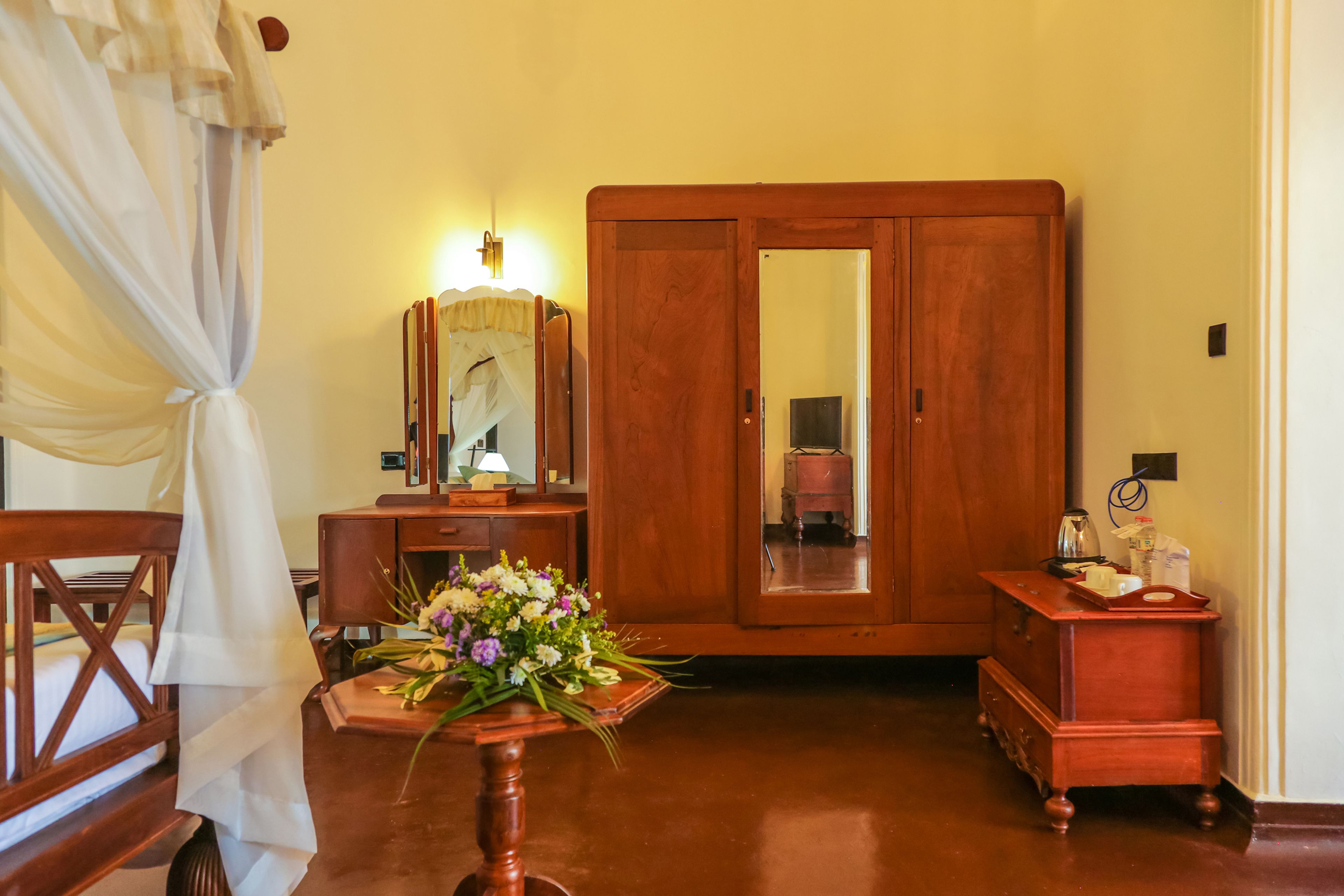 A room with a wooden wardrobe, a small wooden table with a vase of flowers, and a bed with a canopy.