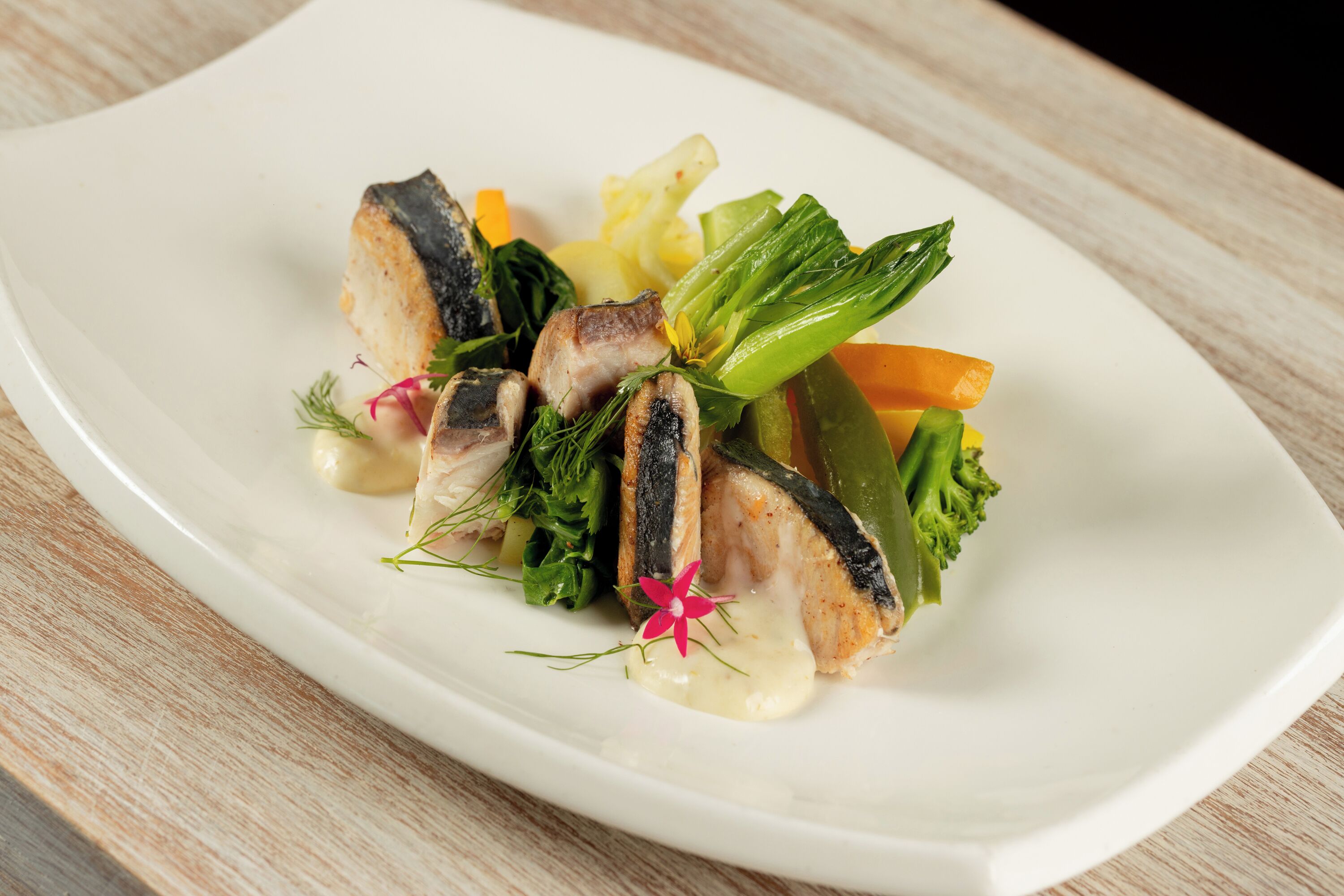 A white plate with a fish dish, vegetables, and a flower garnish.