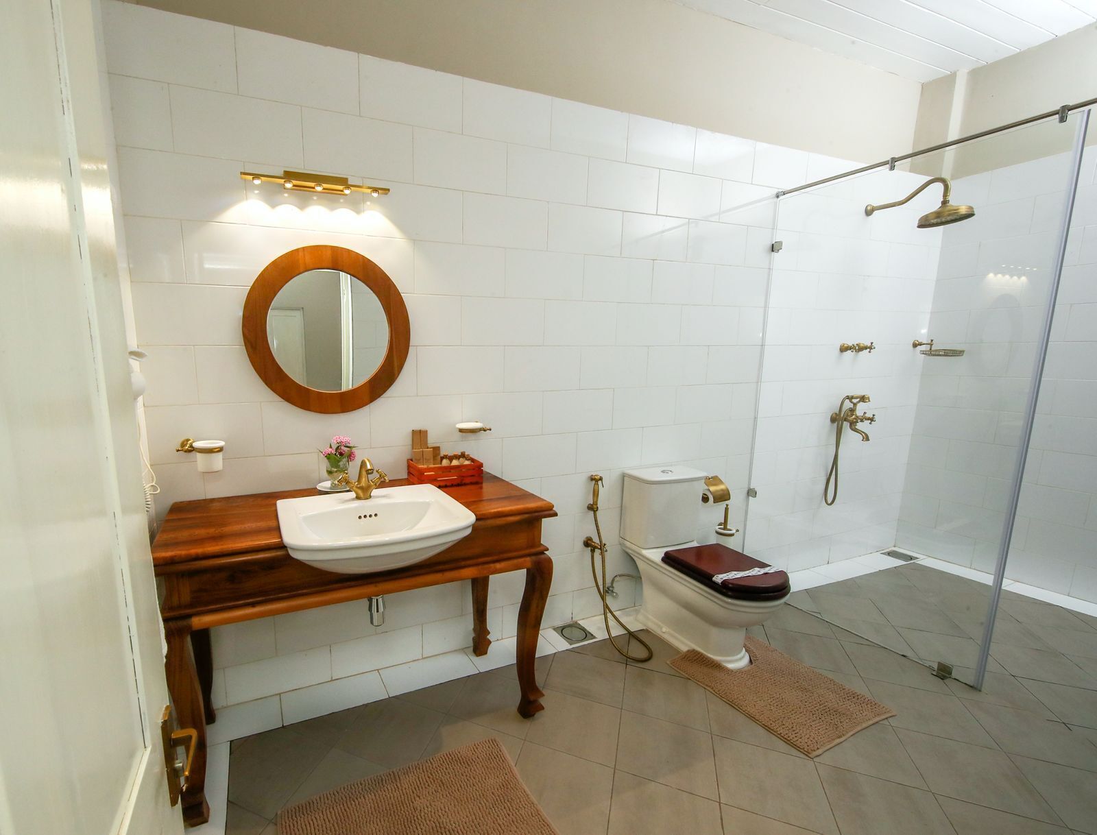 A bathroom with a wooden vanity, a round mirror, a white sink, a toilet, and a glass-enclosed shower area.