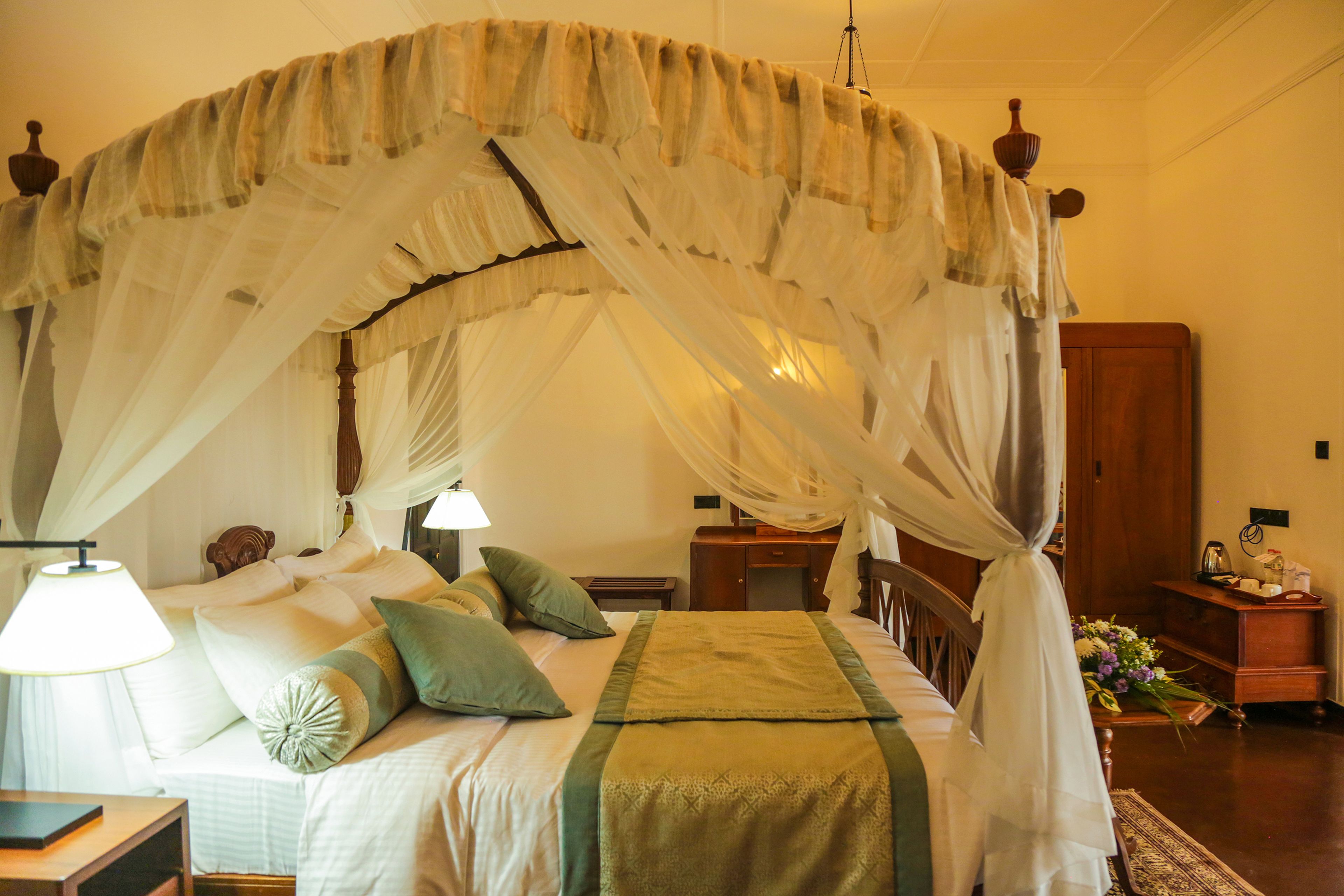 Un lit à baldaquin à quatre poteaux, avec une couvre-lit et des coussins verts, une table de chevet avec une lampe, et une table de nuit en bois ornée d’un vase de fleurs.