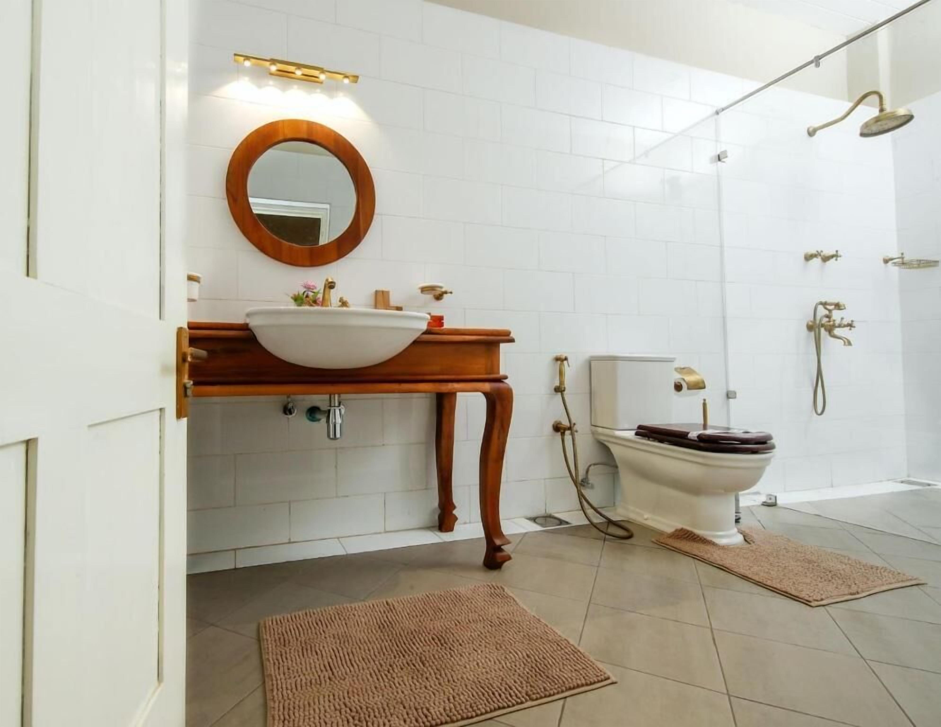 A bathroom with a wooden vanity, a round mirror, a white sink, a toilet, and a shower area.