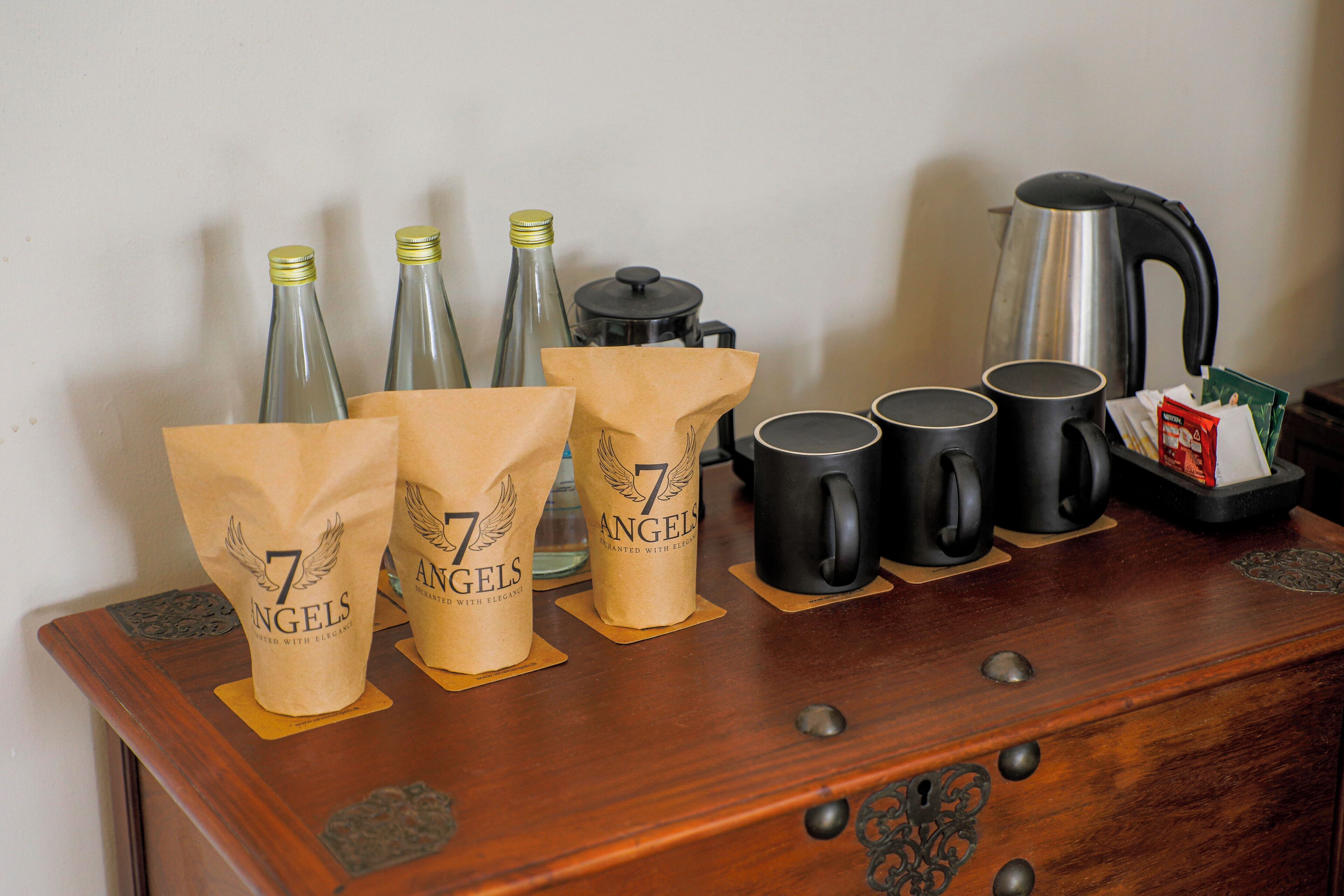 A wooden dresser with decorative metal handles, a coffee maker, four black mugs, and three paper bags with the logo "7 Angels".