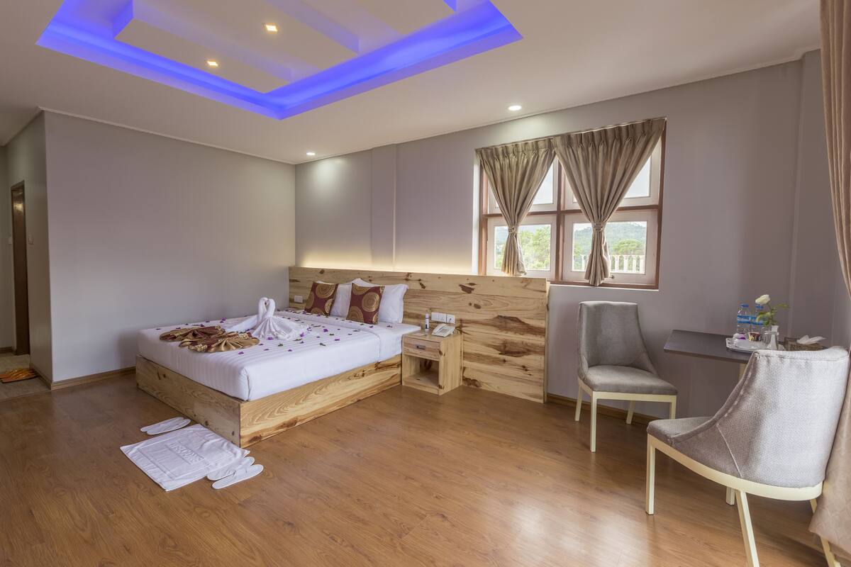 A hotel room with a wooden headboard, a bed with towels, a desk with a chair, and a window with curtains.