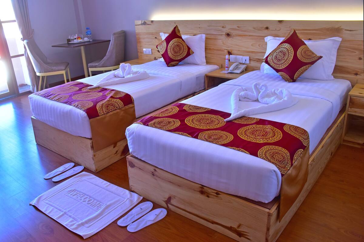 A hotel room with two beds, each with a red patterned bedspread and white pillows. There are two chairs, a small table with a water bottle, and a wooden headboard.