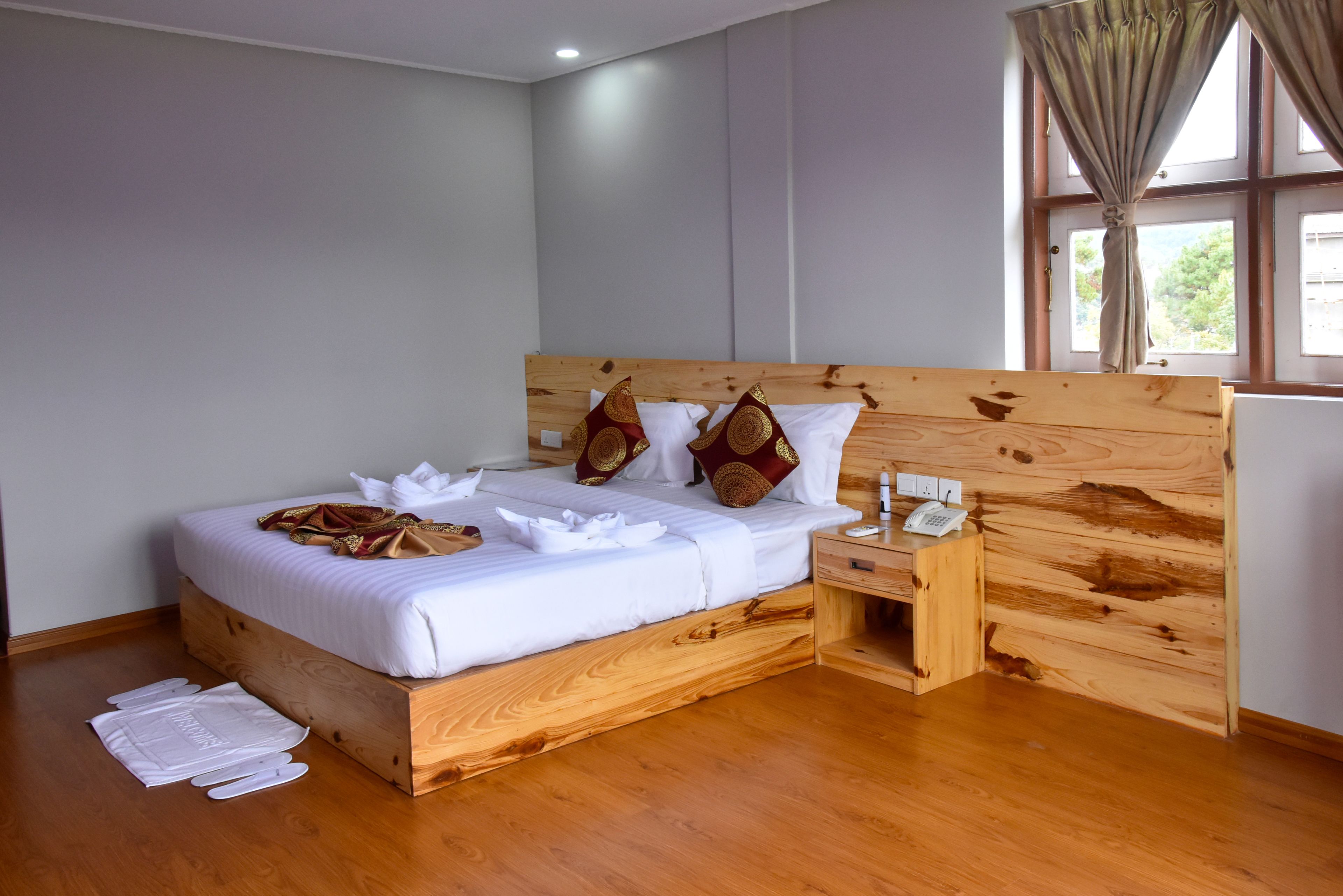 A neatly made bed with white linens and decorative pillows, a wooden headboard, a bedside table with a phone, and a window with curtains.