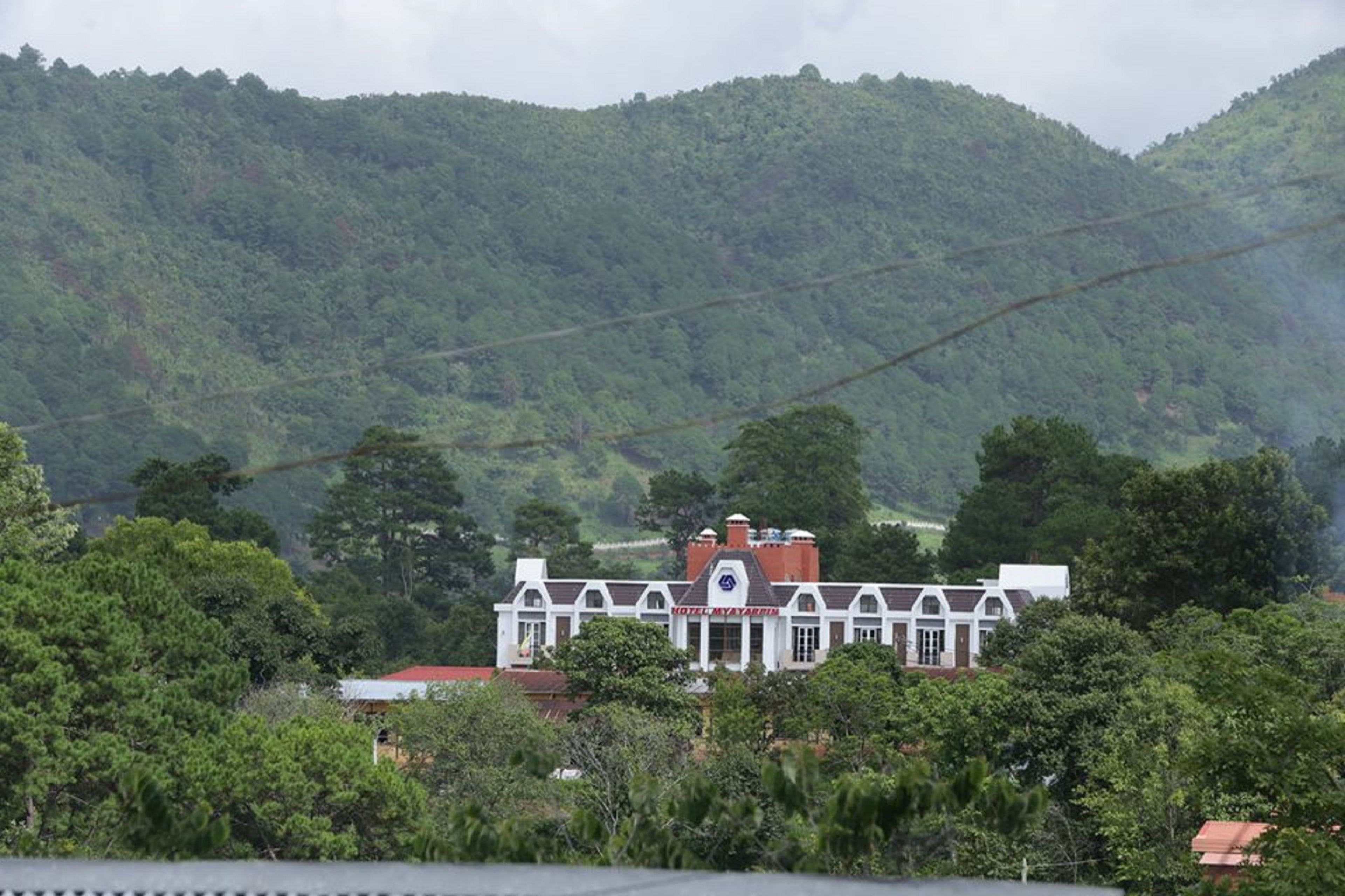 A hotel with a prominent red roof and white walls, surrounded by lush green hills.