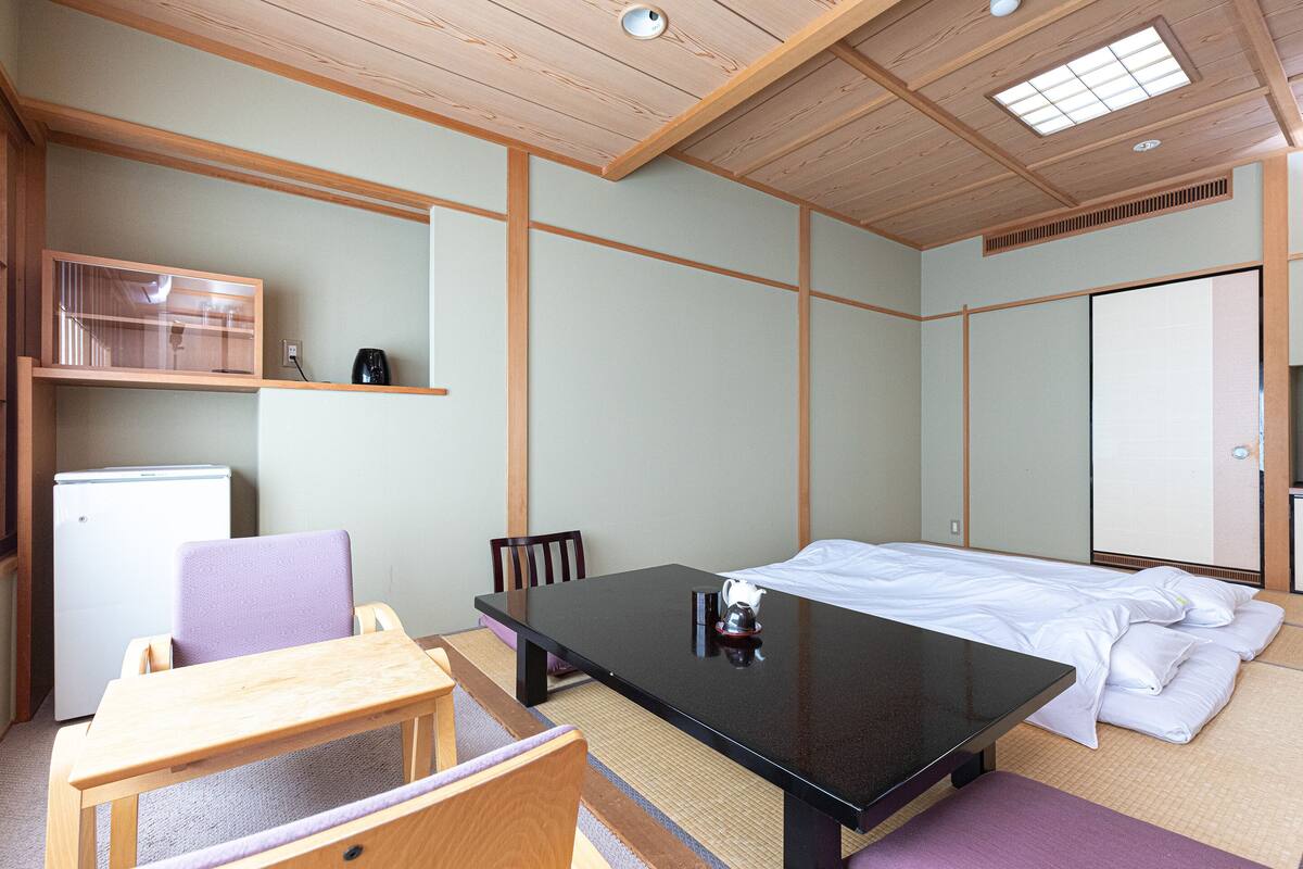 A room with a bed, a table, chairs, a small refrigerator, and a shelf with a kettle.