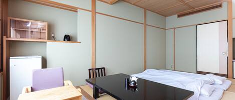 A room with a bed, a table, chairs, a small refrigerator, and a shelf with a kettle.