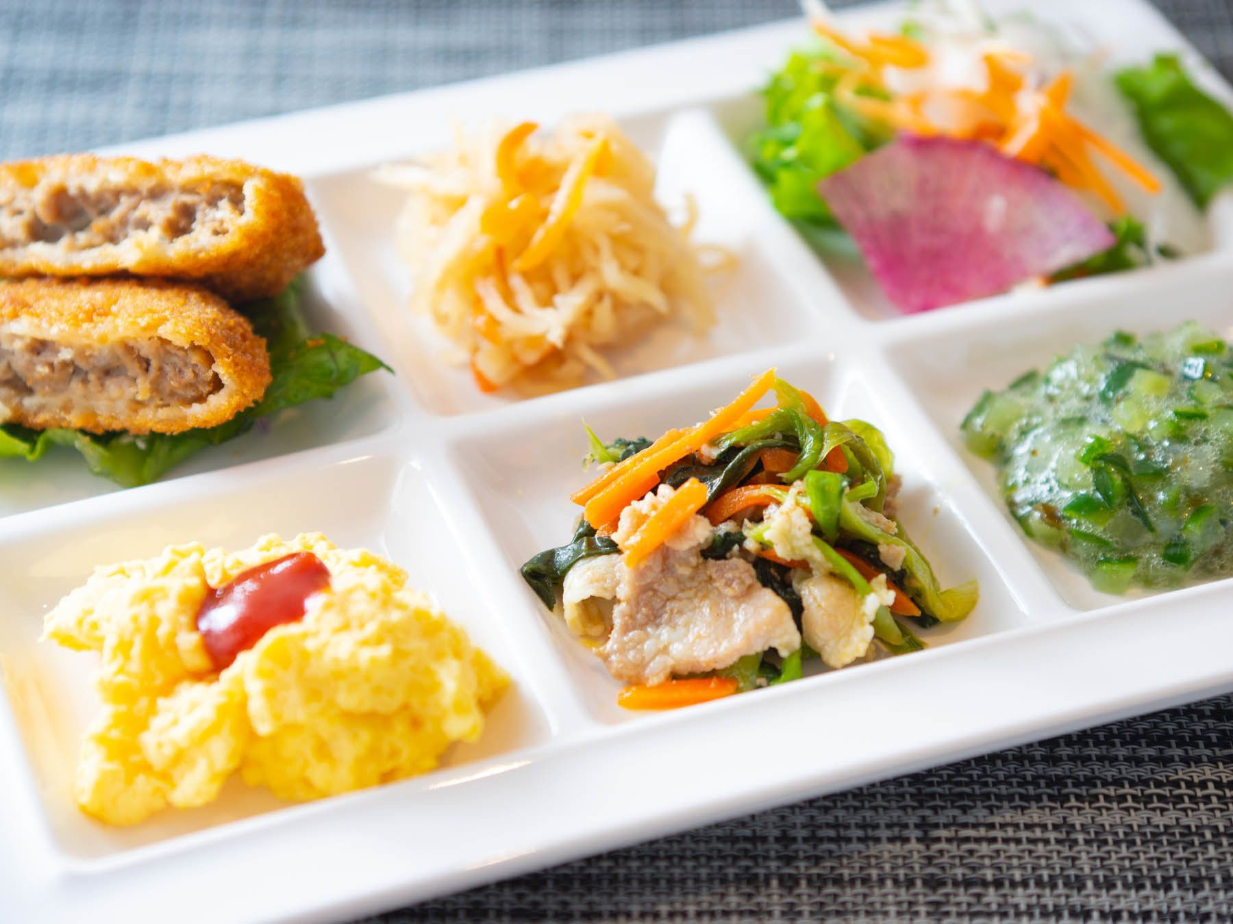 A white rectangular plate divided into sections containing scrambled eggs, a meat dish, shredded cheese, a vegetable salad, and a green sauce.
