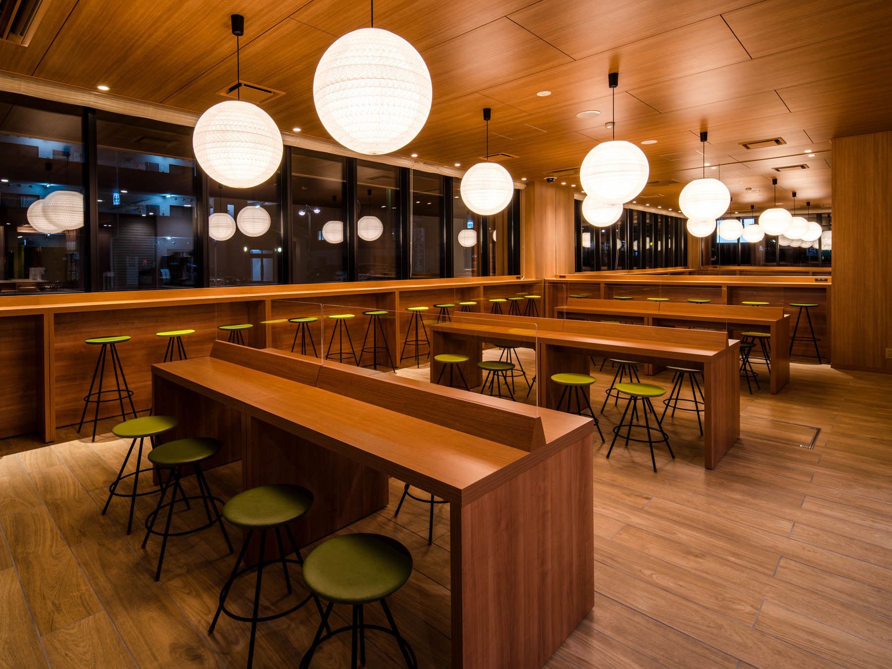 Un interior de restaurante moderno con mesas de madera y taburetes verdes, grandes lámparas colgantes esféricas y vistas de la ciudad a través de amplias ventanas.