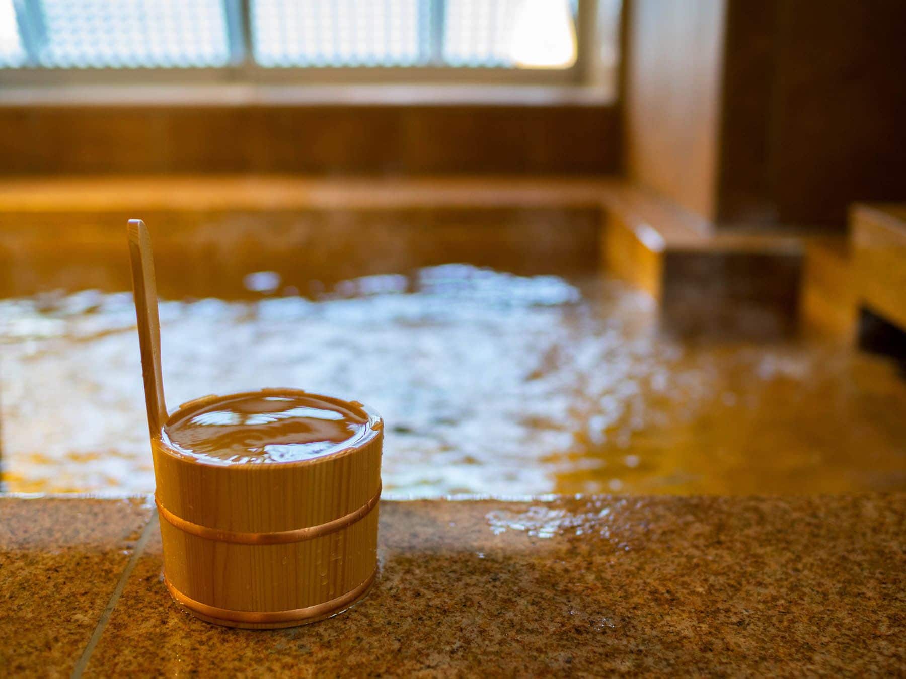 A small wooden container with a straw on a stone surface, likely containing a beverage, in a room with a large window and a tiled floor.
