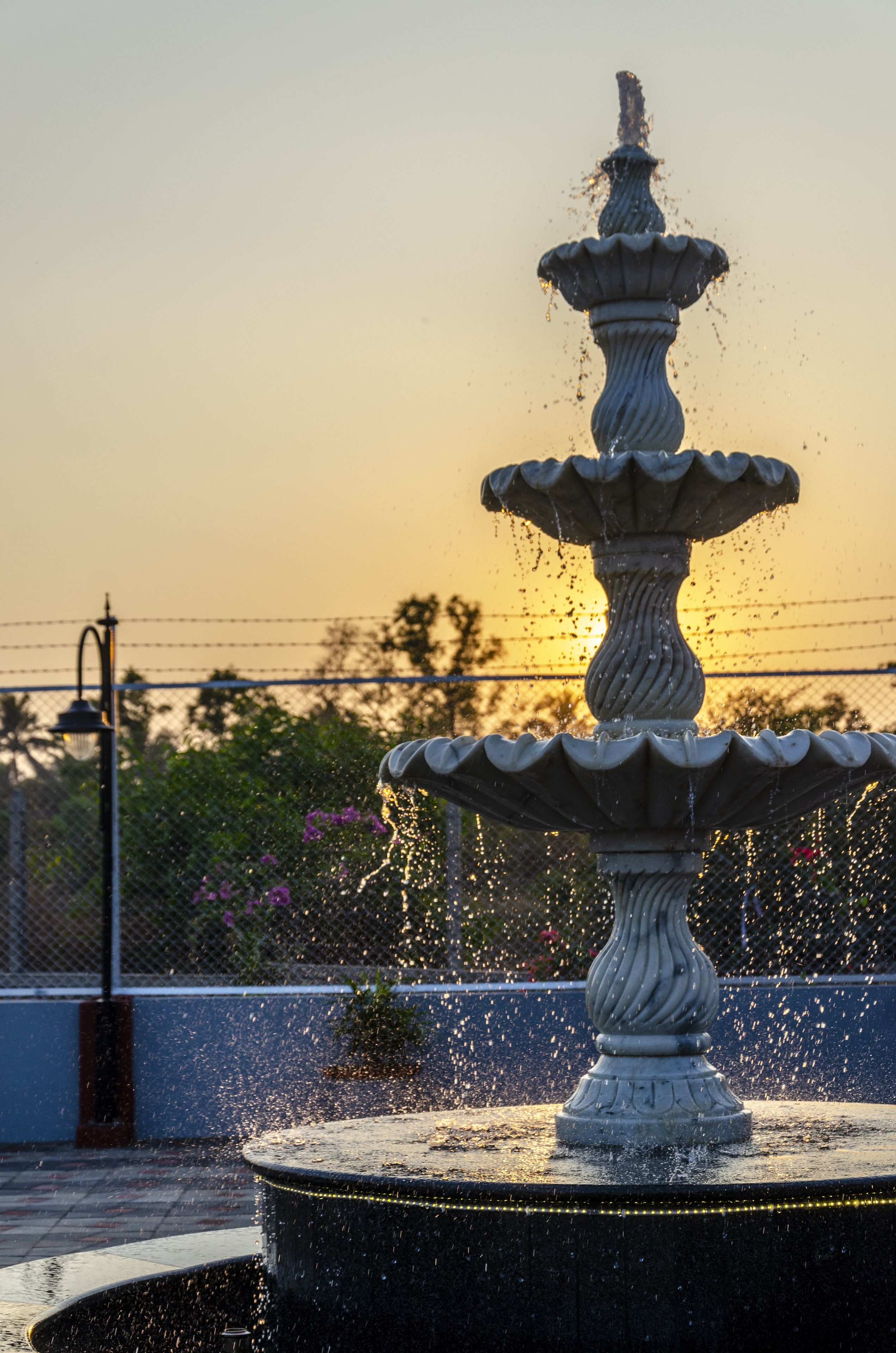 Une fontaine à plusieurs niveaux, agrémentée de jets d’eau, entourée d’une clôture et de plantes, le tout se détachant sur un ciel au coucher du soleil.