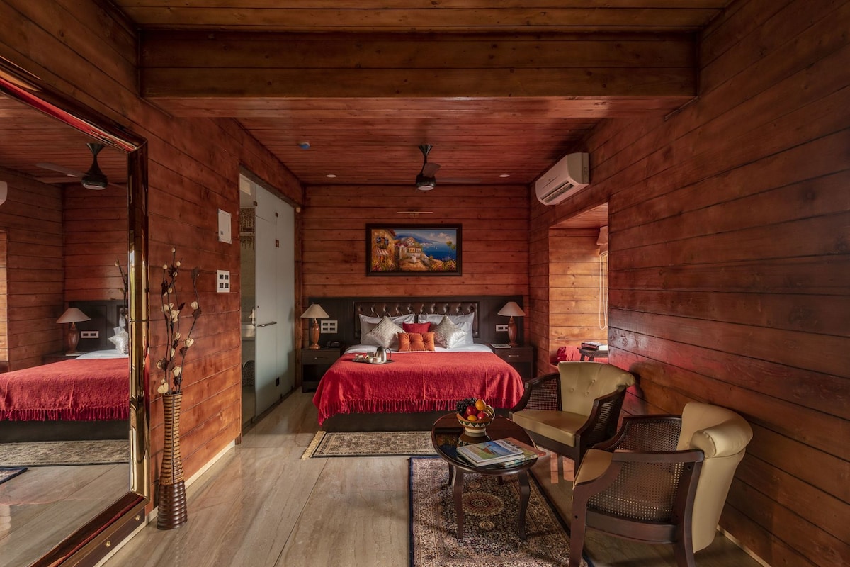 A wooden cabin bedroom with a bed, two chairs, a small table, a mirror, and a painting on the wall.