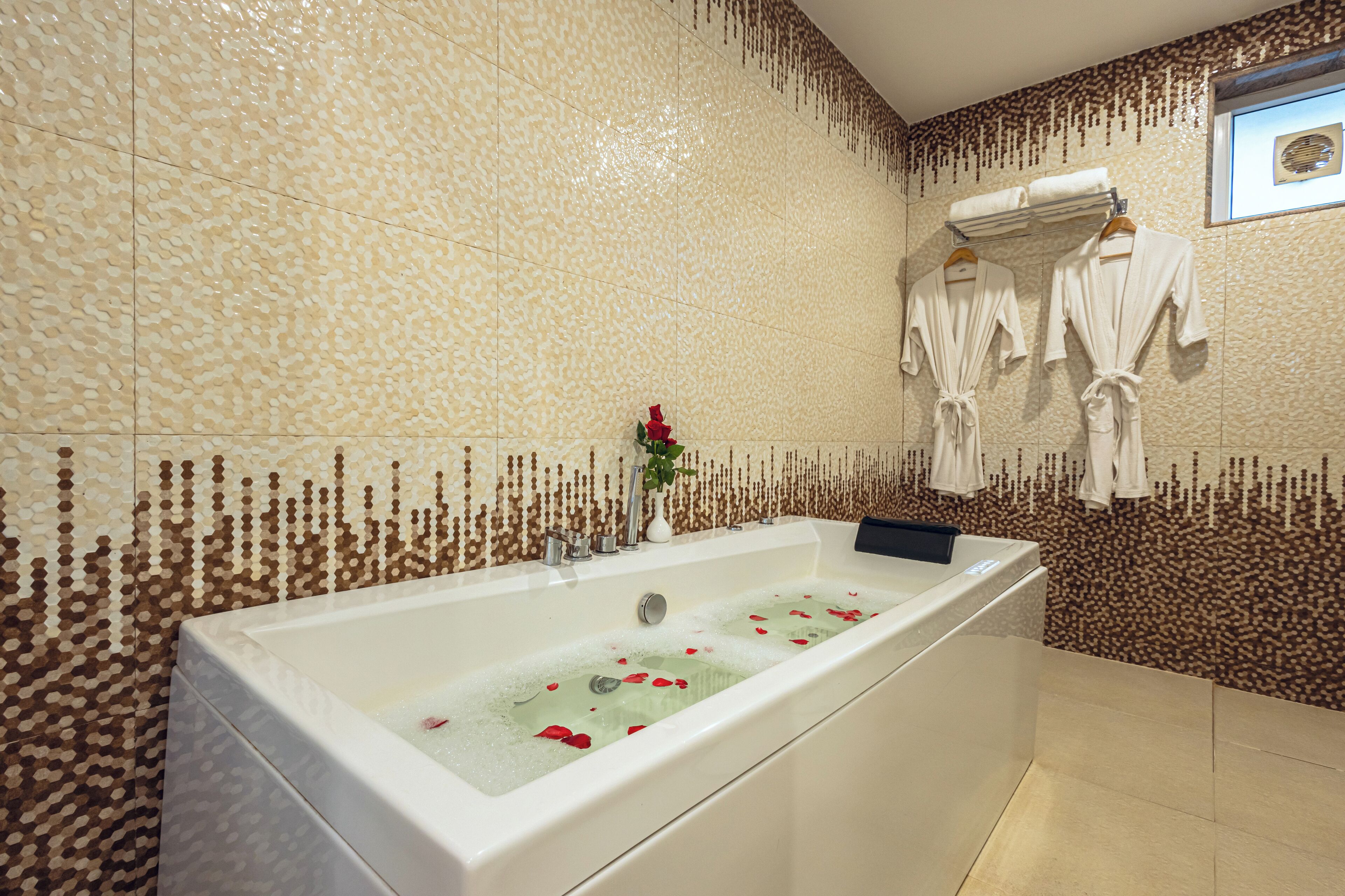 A large bathtub with rose petals in the water, surrounded by beige patterned tiles.