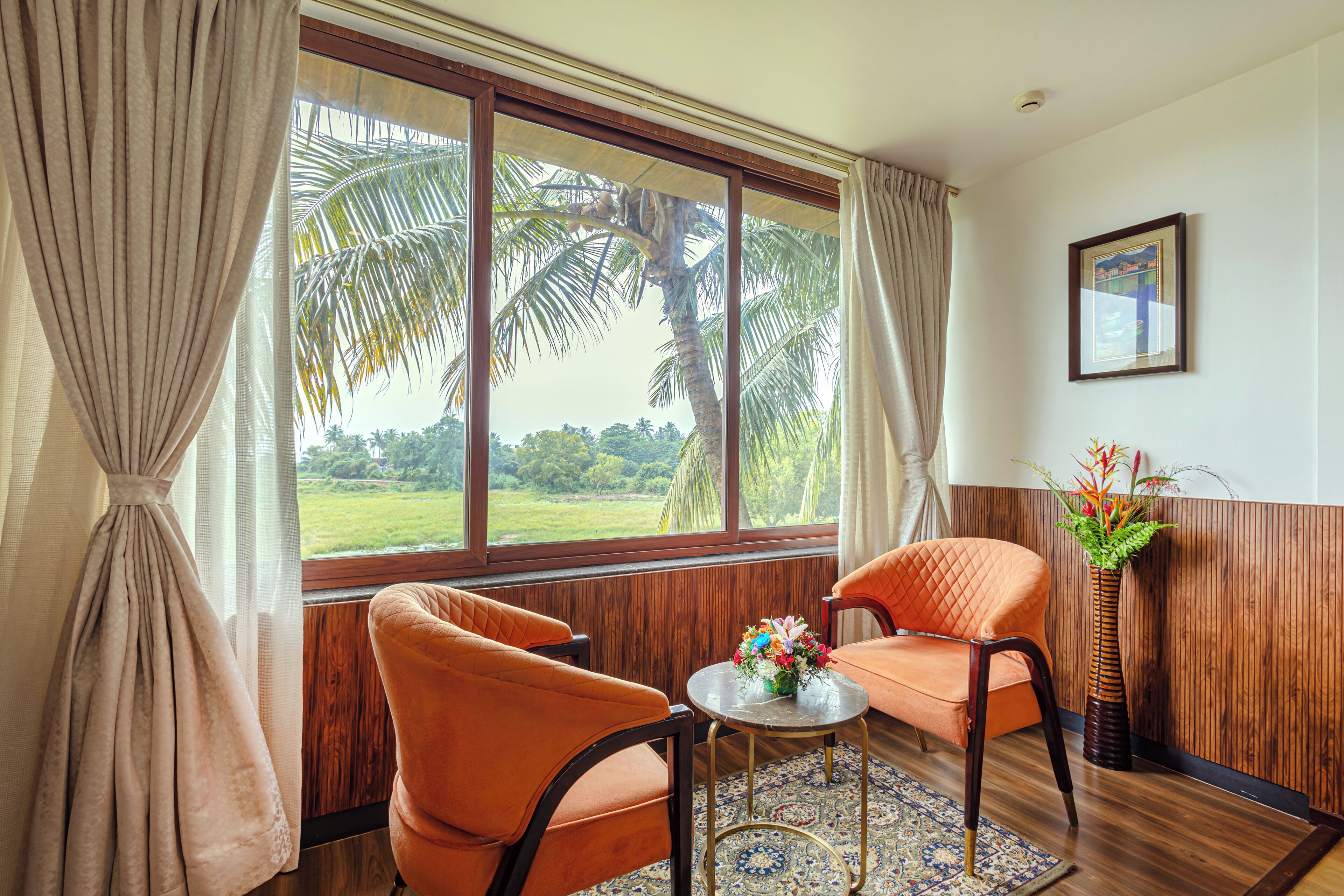 A room with two orange chairs, a small table with a vase of flowers, and a large window with a view of palm trees and greenery.