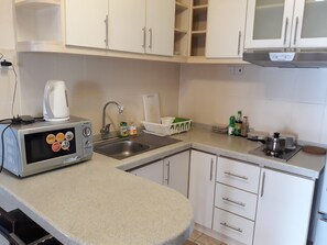 Una cocina con armarios blancos, un lavamanos de acero inoxidable, una tetera, un microondas y varios utensilios de cocina sobre la encimera.