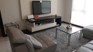 Una sala de estar moderna con un sofá gris, una mesa de centro de vidrio, un televisor de pantalla plana sobre un mueble blanco y una alfombra gris y esponjosa.