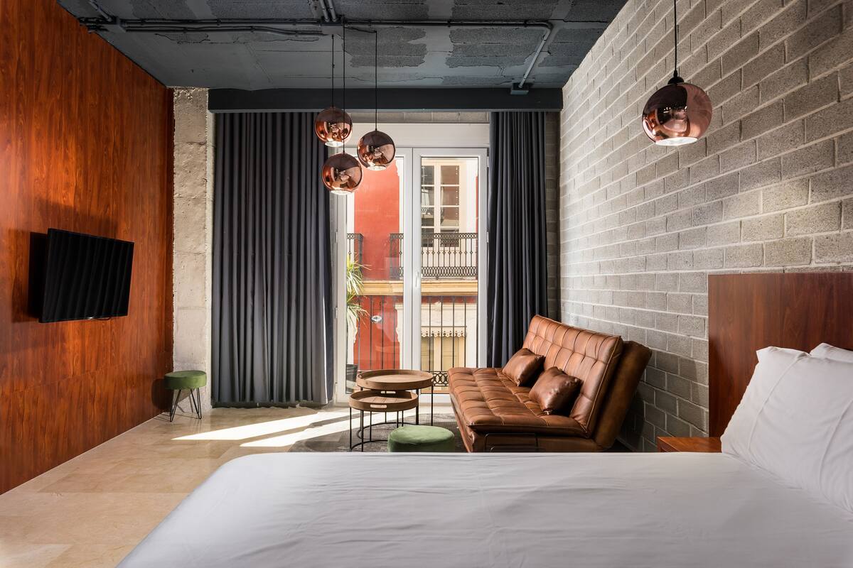 Una habitación de hotel moderna con una cama grande, un sofá de cuero, una pequeña mesa redonda y un televisor montado en la pared.