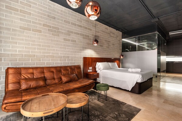 A modern bedroom with a brick wall, a leather sofa, a wooden coffee table, a green ottoman, and a bed with white linens.