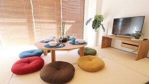 A modern living room with tatami flooring, a round table with dinnerware, colorful cushions, a television, and a potted plant. - Mainichi Building Osaka (Osaka)