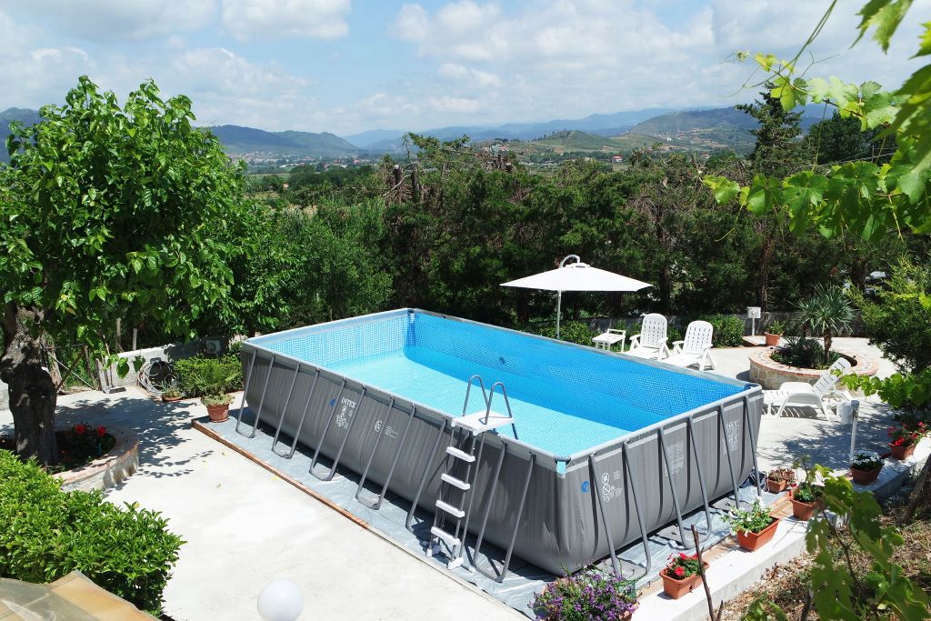 Una piscina elevada con escalera, rodeada de plantas en macetas y muebles de exterior, con montañas y vegetación de fondo.