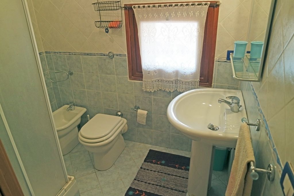 A bathroom with a toilet, bidet, sink, and a window with a lace curtain.
