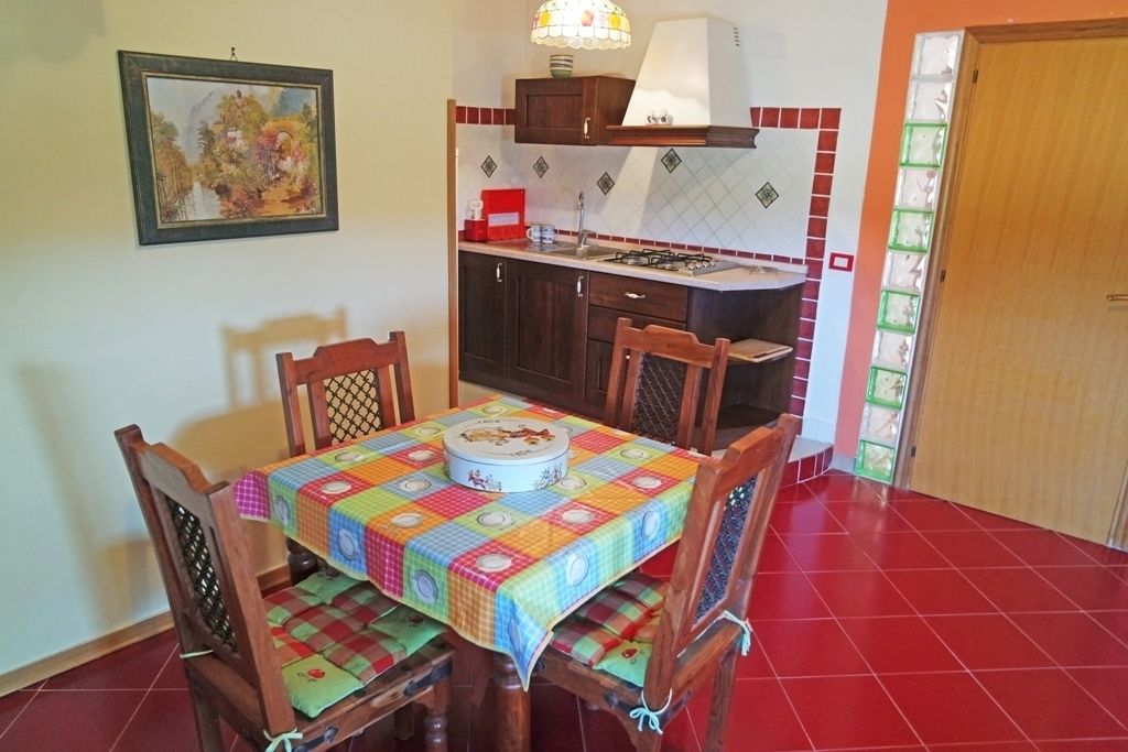 A dining area with a table covered in a colorful tablecloth, surrounded by wooden chairs with cushioned seats. A painting hangs on the wall above the table.
