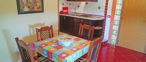 Un comedor con una mesa cubierta por un mantel colorido, rodeada de sillas de madera con asientos acolchados. Un cuadro cuelga en la pared sobre la mesa.