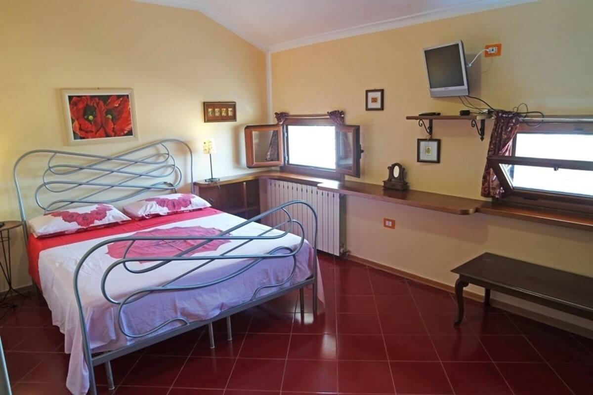 Une chambre à coucher avec un lit en métal, une fenêtre avec des rideaux, une table de chevet et un téléviseur fixé au mur.