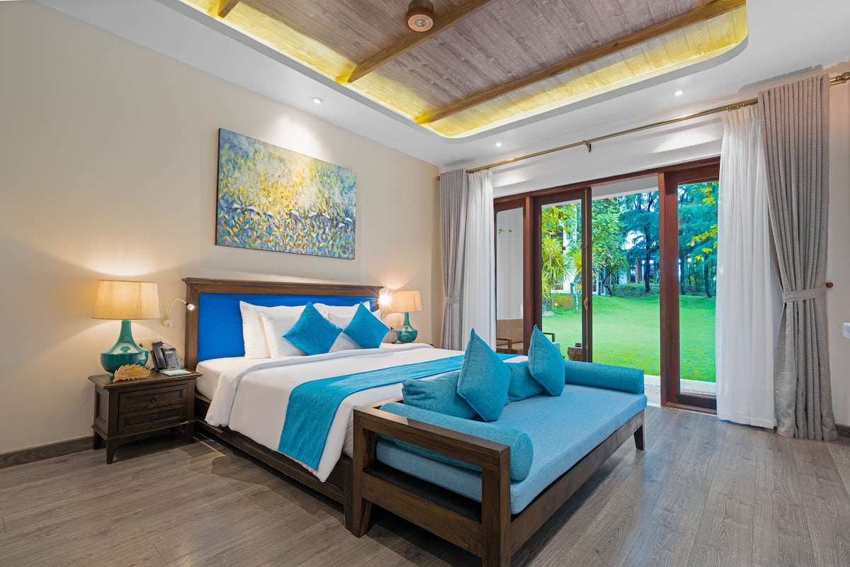 A spacious bedroom with a large bed, a nightstand, and a view of a lush garden through a sliding glass door.