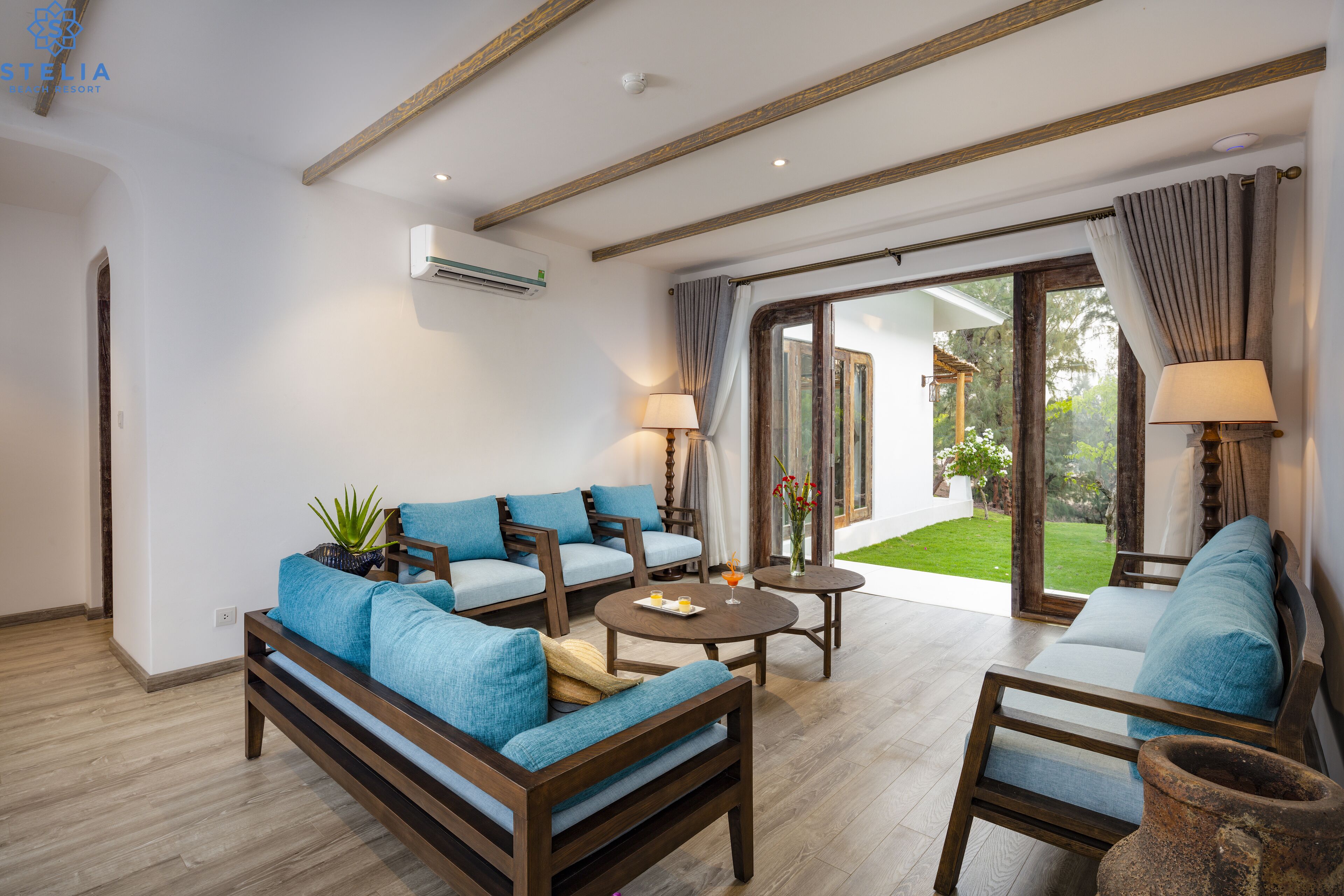 A spacious living area with a sofa set, two armchairs, a coffee table, and a large sliding glass door leading to a garden.