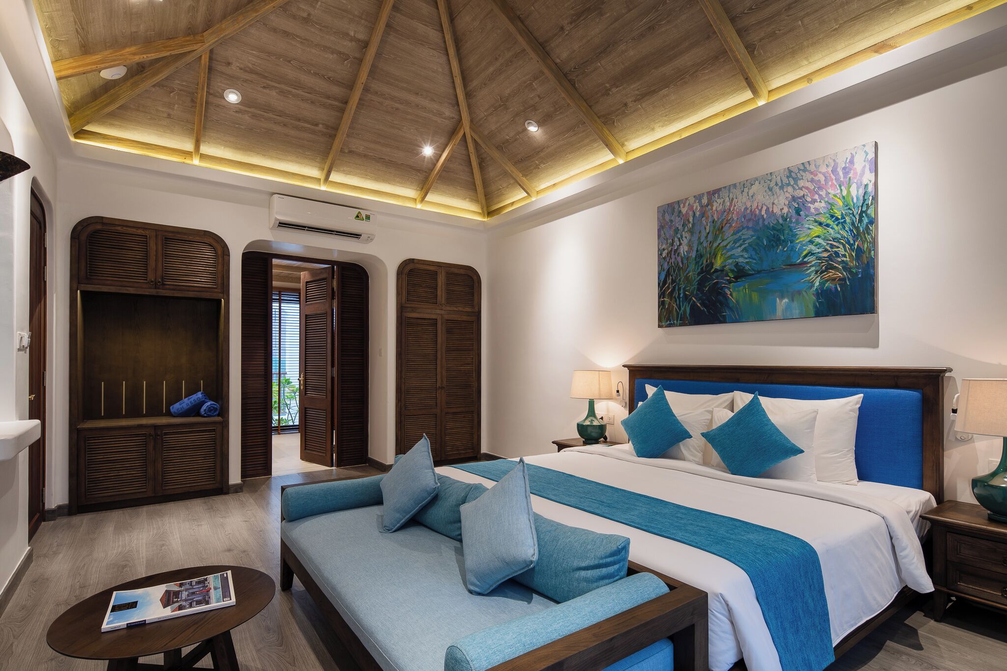 Una habitación de hotel con techo de madera, una cama grande con ropa de cama azul, un sofá con cojines azules, un cuadro en la pared y una ventana con contraventanas.