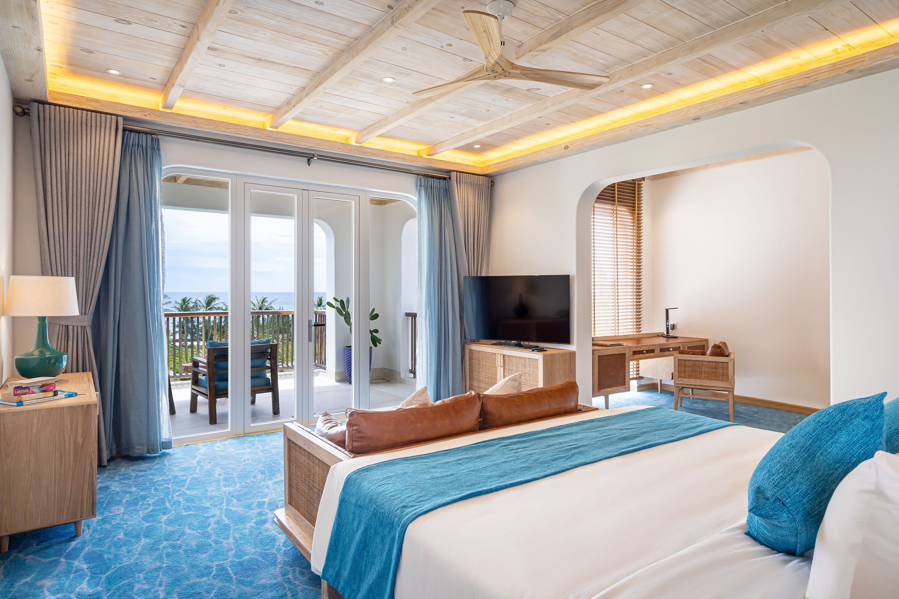 A hotel room with a large bed, a desk with a TV, and a balcony with a view.