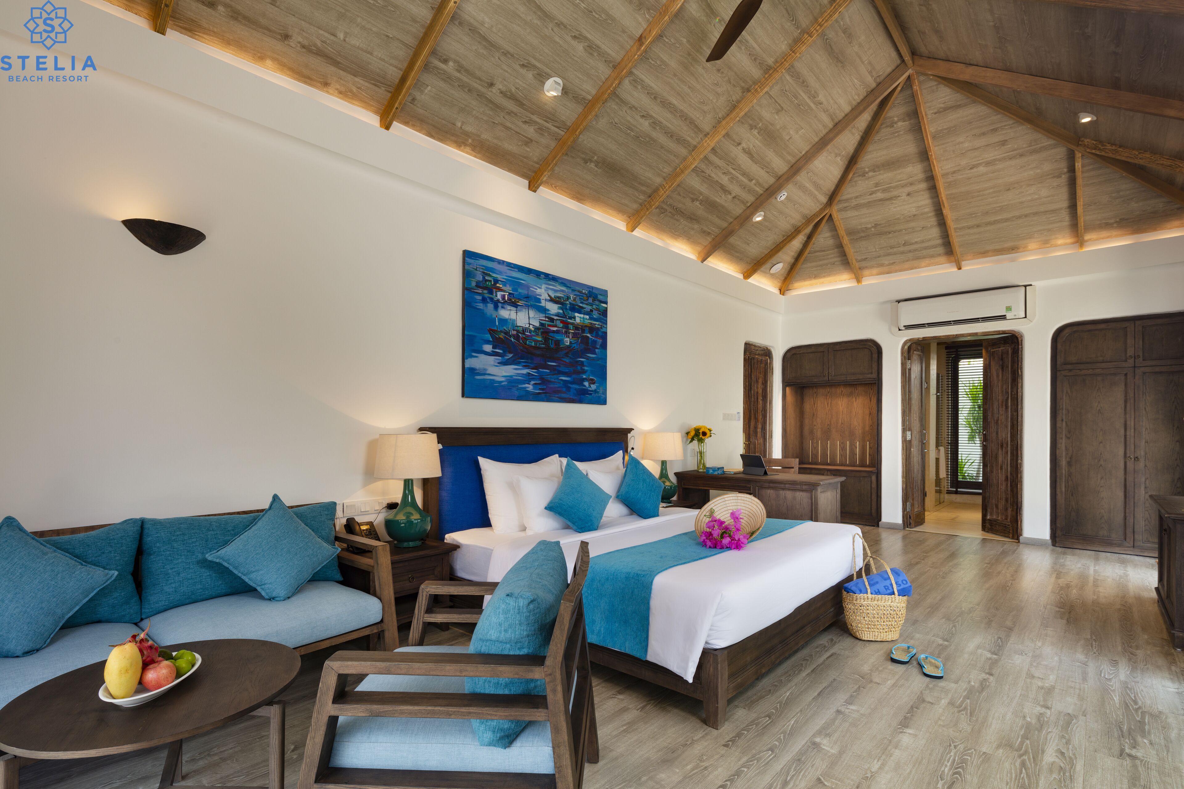A spacious bedroom with a large bed, a sofa, and a small table. There is a painting on the wall and a wooden ceiling.
