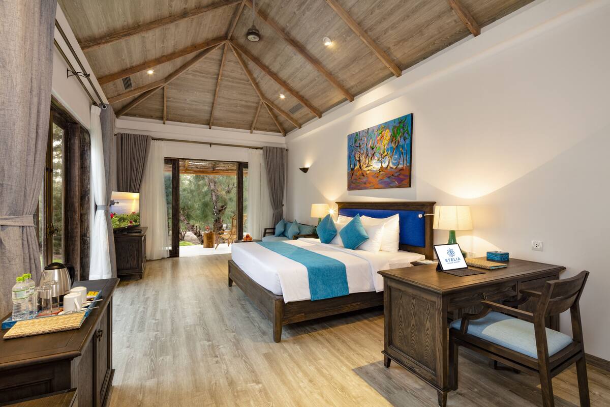 A bedroom with a wooden ceiling, a bed with blue and white bedding, a desk with a laptop, and a painting on the wall.
