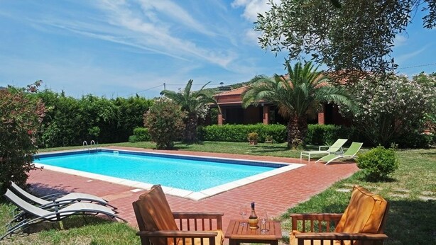 A swimming pool with lounge chairs and a table, surrounded by greenery and a house in the background. - Villa Kalithea (Montecorice)