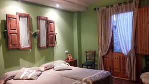 Una habitació amb dos llits, una porta de fusta, una cadira i una finestra amb cortines.