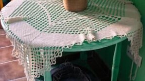 Una taula rodona amb un mantell de ganxet, un vas verd amb flors blanques, i una cistella a sota.