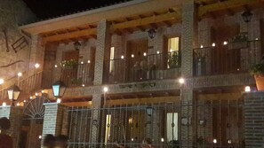 A multi-story building with brick walls and wooden beams, featuring balconies with hanging lights and potted plants. - Alojamientos Rurales La Aldaba (Navalagamella)