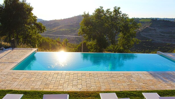 A rectangular swimming pool with a clear blue water surface, surrounded by a stone patio and a row of white lounge chairs. - Casale Acquaviva (Torchiara)