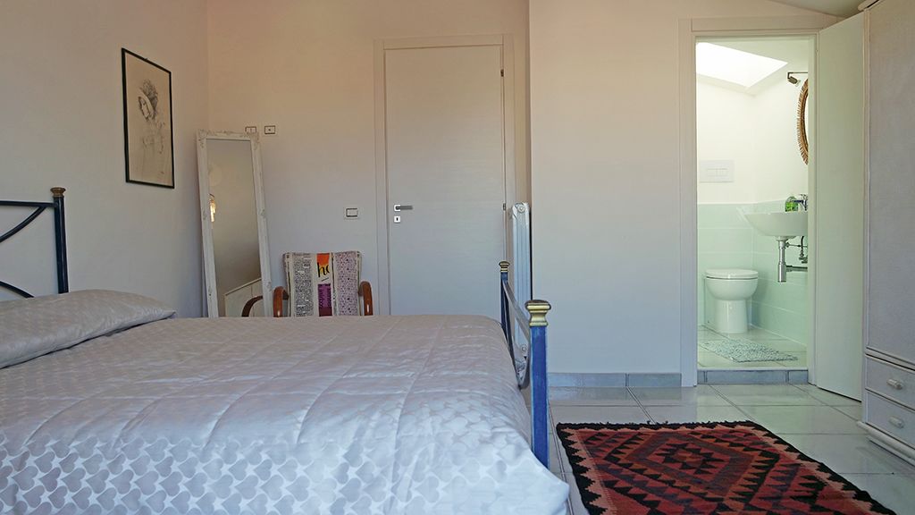 A bedroom with a bed, a bedside table, a mirror, and a bathroom visible through an open door.